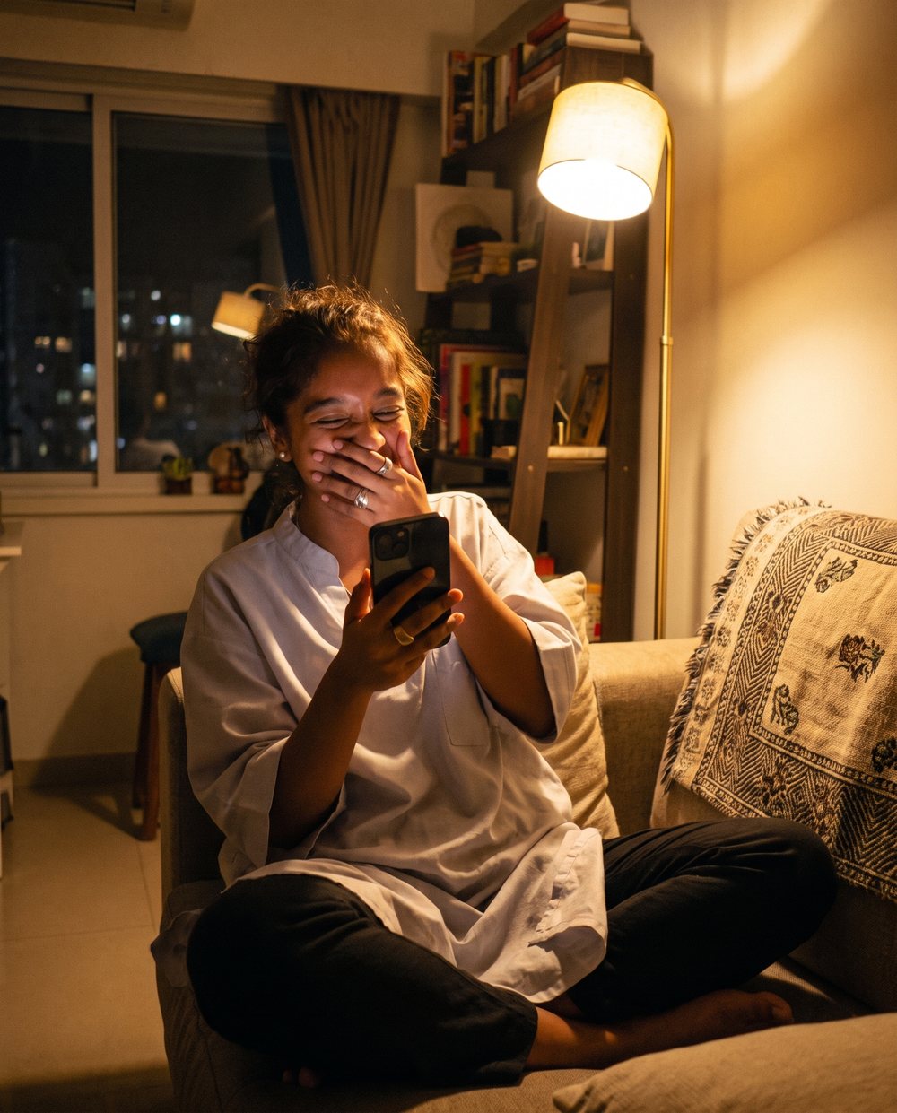 A woman on a sofa laughing behind her hand at something on her phone
