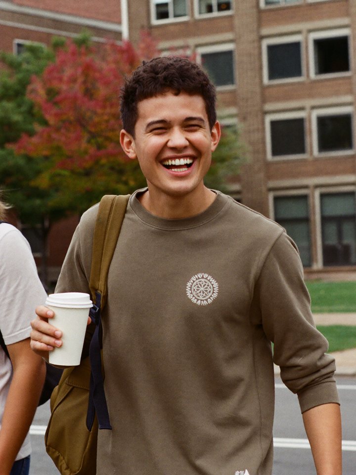 A student walking across campus in autumn with a coffee, laughing