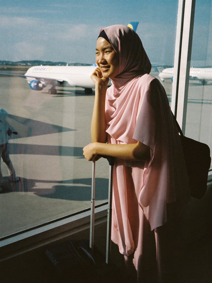 A woman in a hijab at an airport window with her suitcase, looking out at a plane