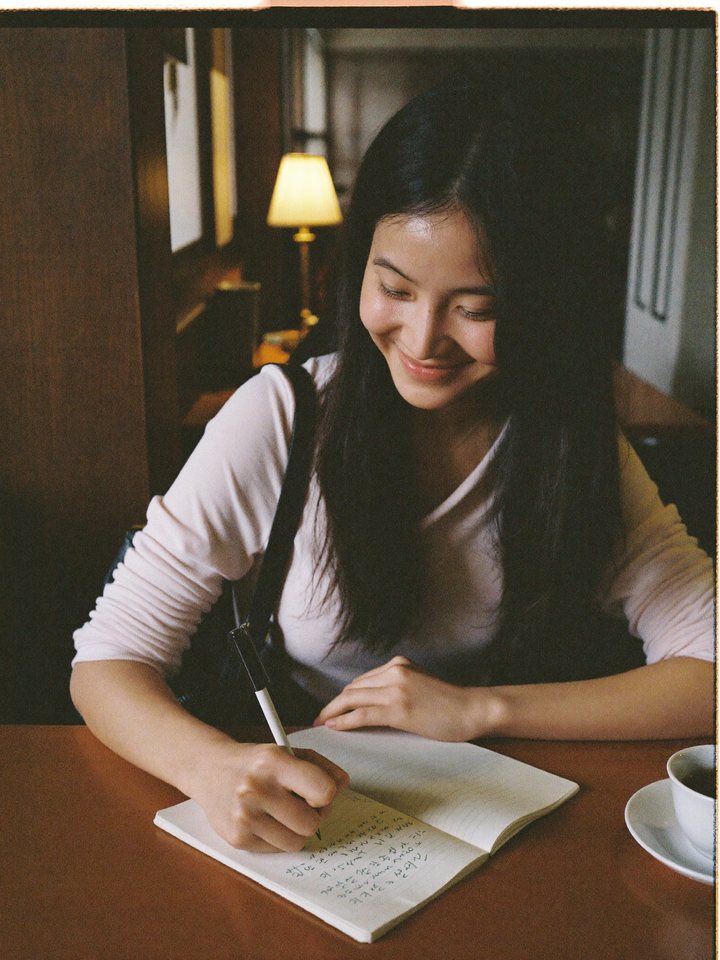 A woman writing in a notebook at a cafe, smiling to herself