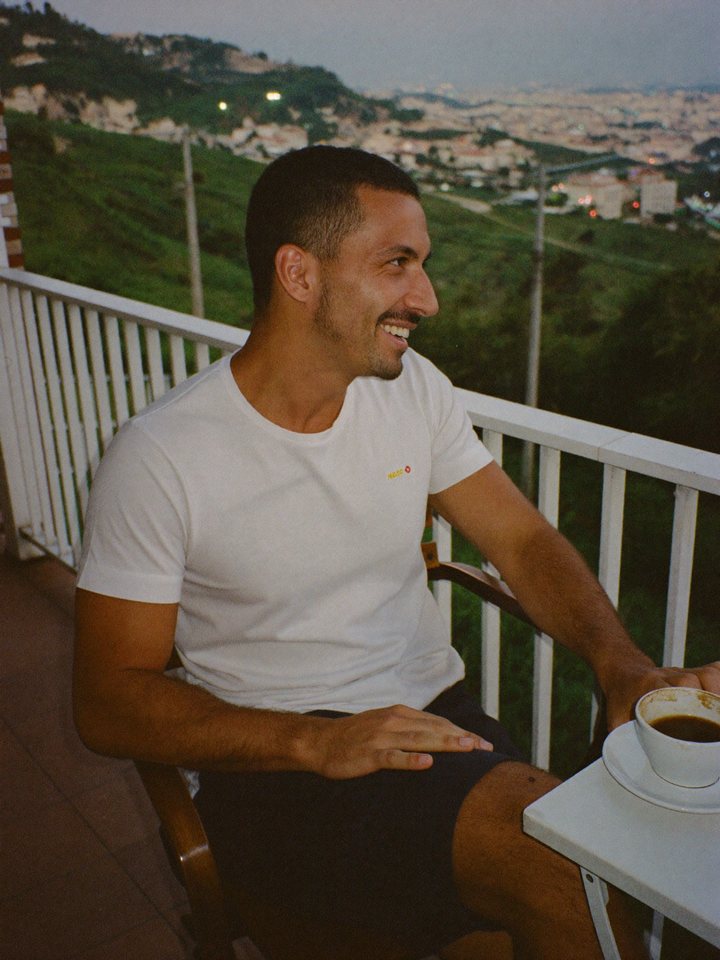 A man laughing softly at a balcony table at dusk, coffee in hand
